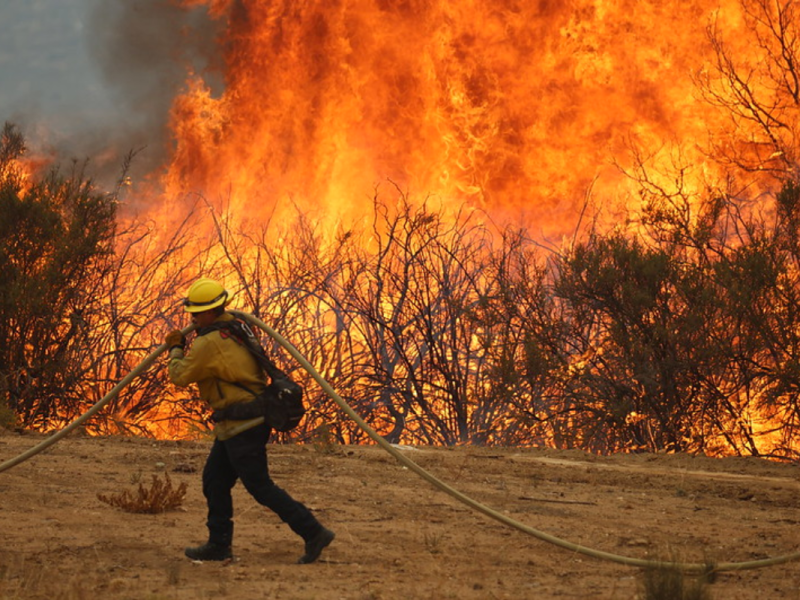 A firefighter carries a hose as flames burn in the background.