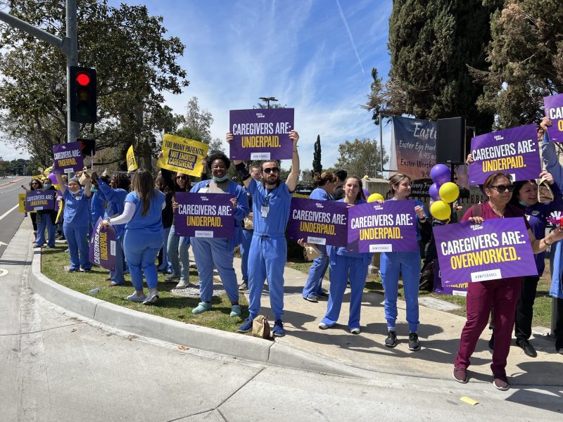 A group of healthcare workers wearing scrubs hold up signs that say, “Too many patients. Not enough caregivers,” “Our hospital is: understaffed,” and “Caregivers are: overworked,” on a street corner at the entrance of Riverside Community Hospital.