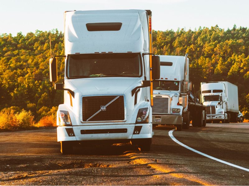 Three semi-trucks driving down a damaged road at sunset.