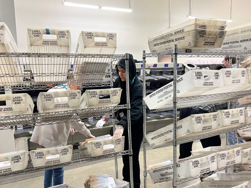 Elections workers stand behind a shelf full of sorted vote-by-mail ballots.