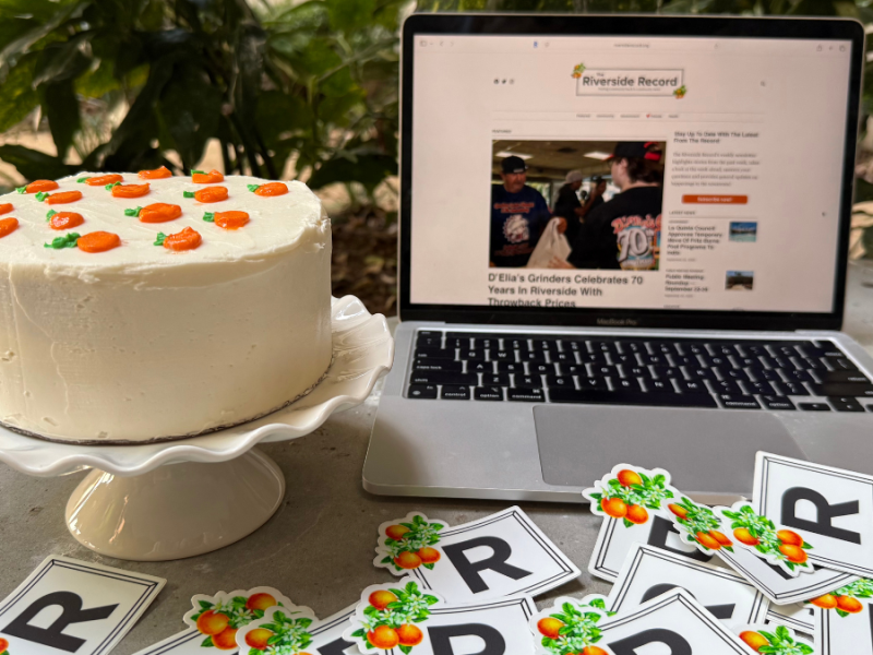 A photo of a cake with orange decorations, Riverside Record stickers and a laptop with The Riverside Record's website pulled up.