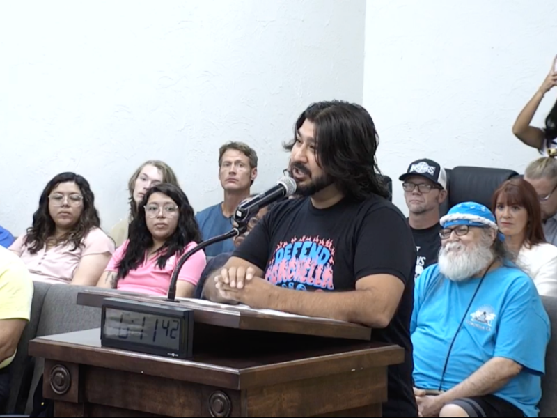 A person wearing a "Defend Coachella" shirt talking into a microphone at a podium.