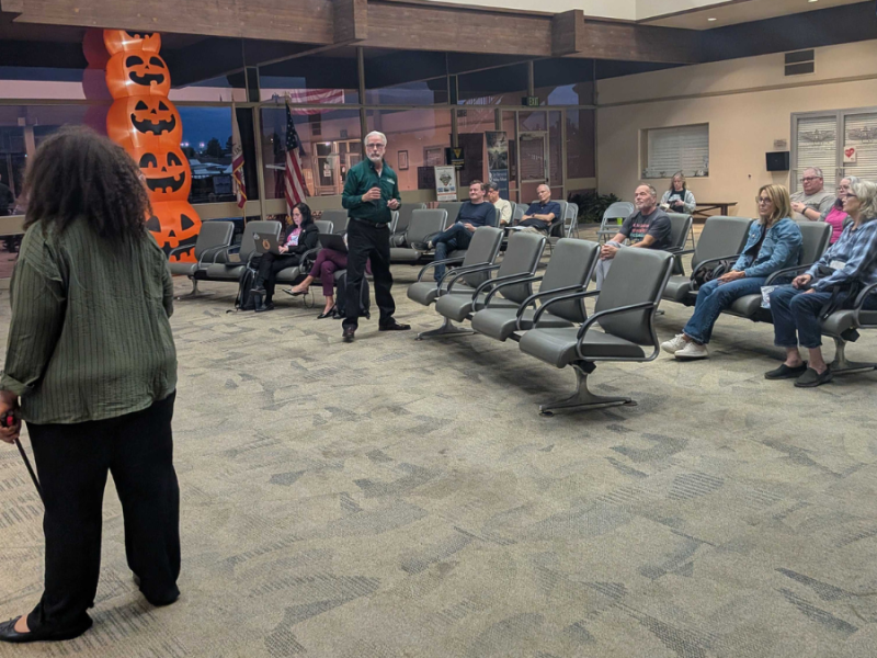 A speaker in a group of people talks to a person taking notes at the front of the room.